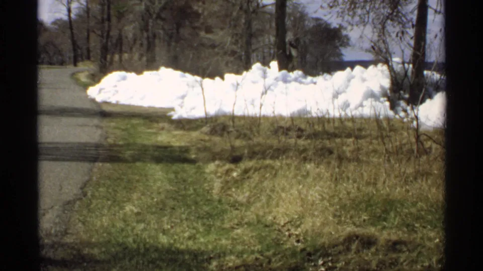 1970 Minnesota 8mm frame looking along a quiet road bordered by melting snow, leafless trees, rough grass, and distant water.