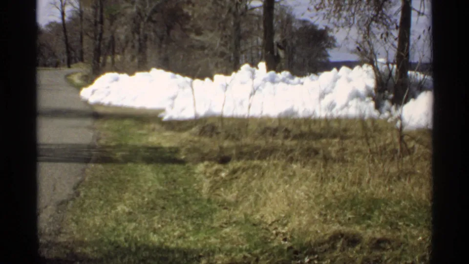 1970 Minnesota 8mm frame of a roadside snowbank receding into bare trees, with grass in the foreground and water beyond.