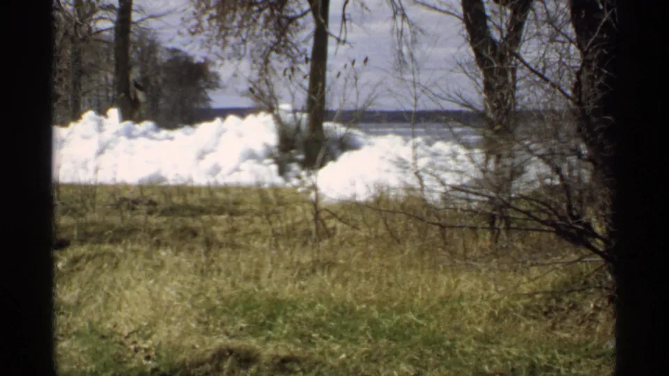 1970 Minnesota home-movie view of bright thawing snow piles, brown-green grass, bare trees, and water in the distance.