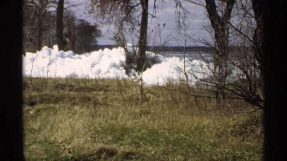 Minnesota 8mm footage frame with melting snowbank, dormant grass, bare trunks, brush, and a distant lakeside horizon.
