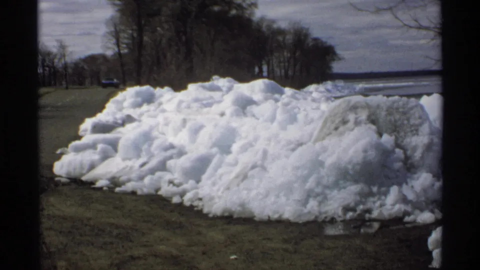 1970 Minnesota frame showing a close roadside pile of dirty melting snow, with trees, pavement, and distant water beyond.