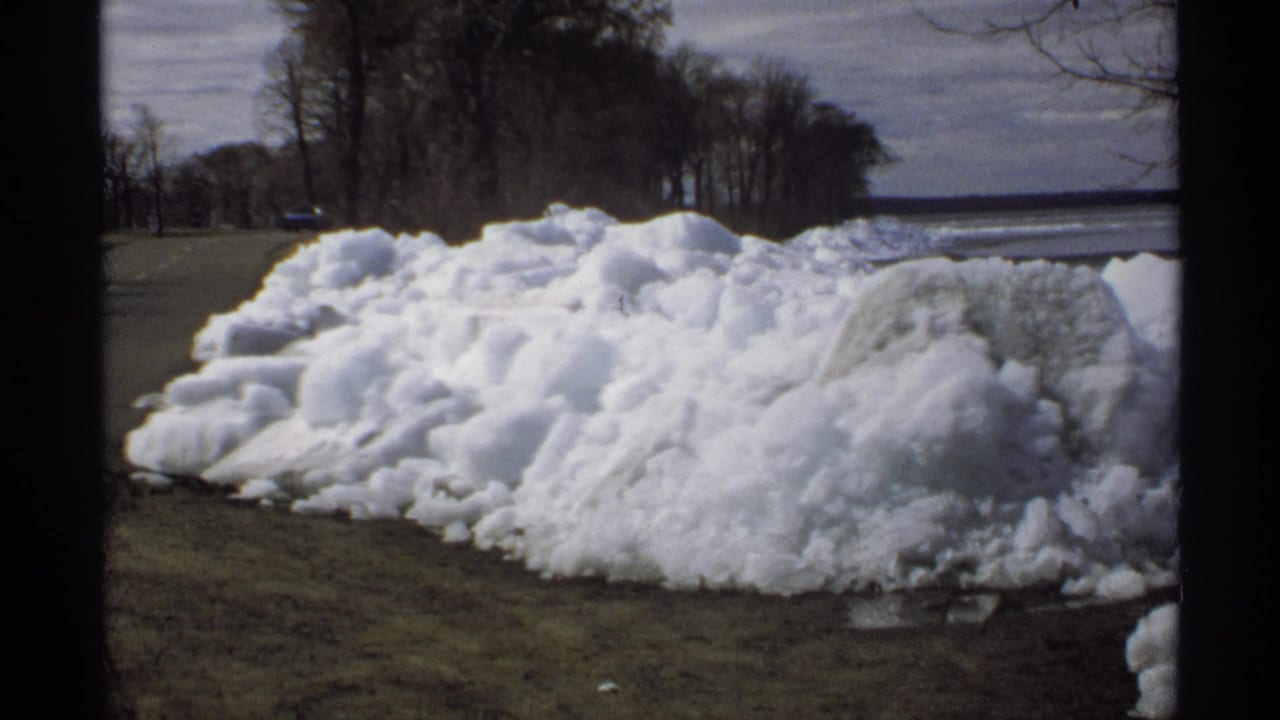 Large melting snow pile beside a road with bare trees and distant water.
