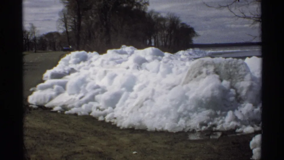 1970 Minnesota 8mm frame showing a lumpy melting snowbank on grass beside a quiet road, with bare trees and water in the distance.