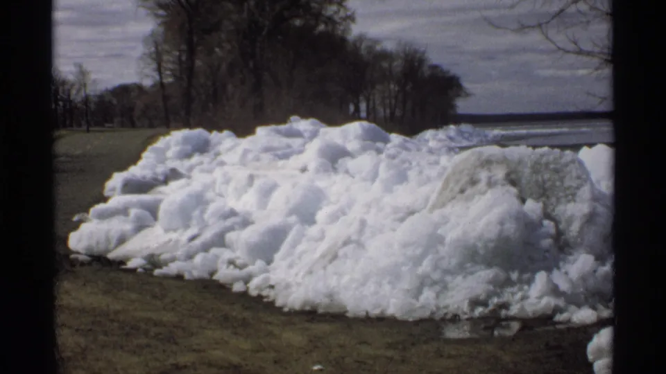 1970 Minnesota home-movie frame of a melting roadside snow pile, with dark tree line and a cold-looking body of water beyond it.