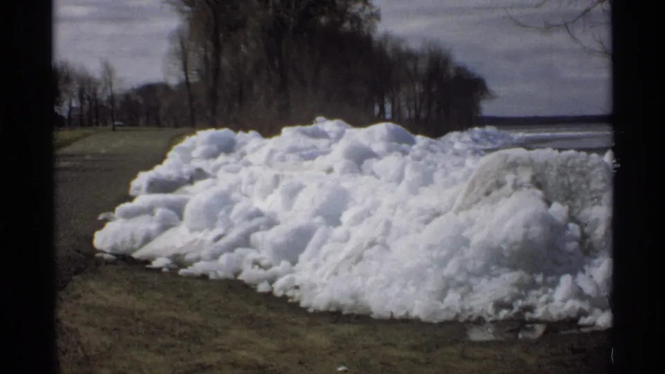 1970 Minnesota 8mm view of a large uneven snow pile thawing along a grassy roadside, with leafless trees behind it.
