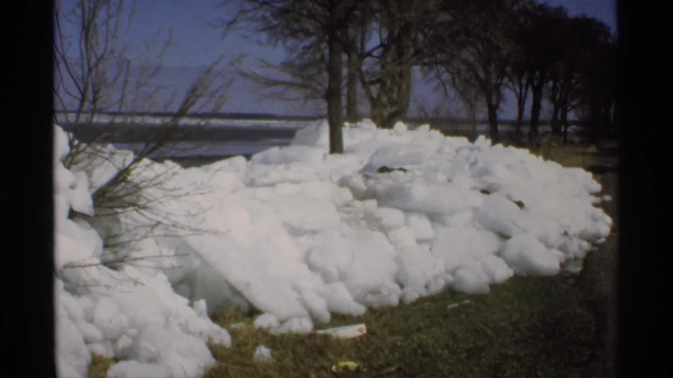 1970 Minnesota frame showing snow-covered grass near bare trees, with a partially visible body of water beyond the shoreline.