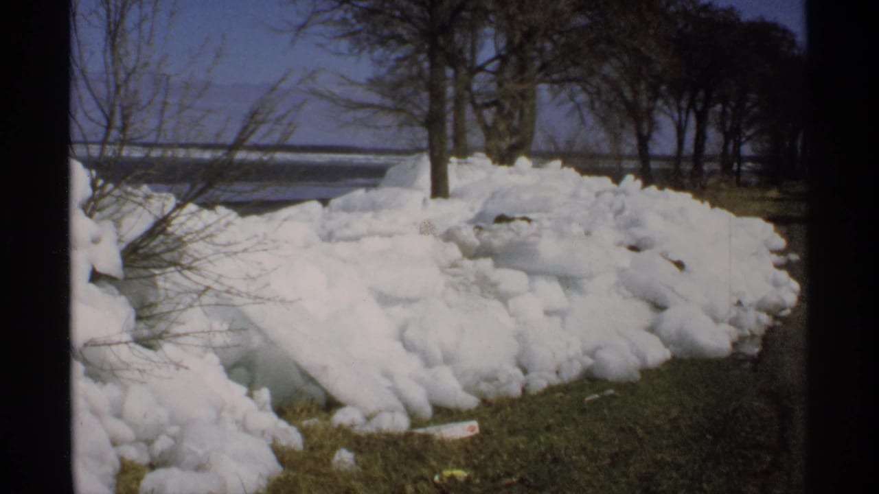 Piled snow beside grass, bare trees, road, and water under a blue sky.