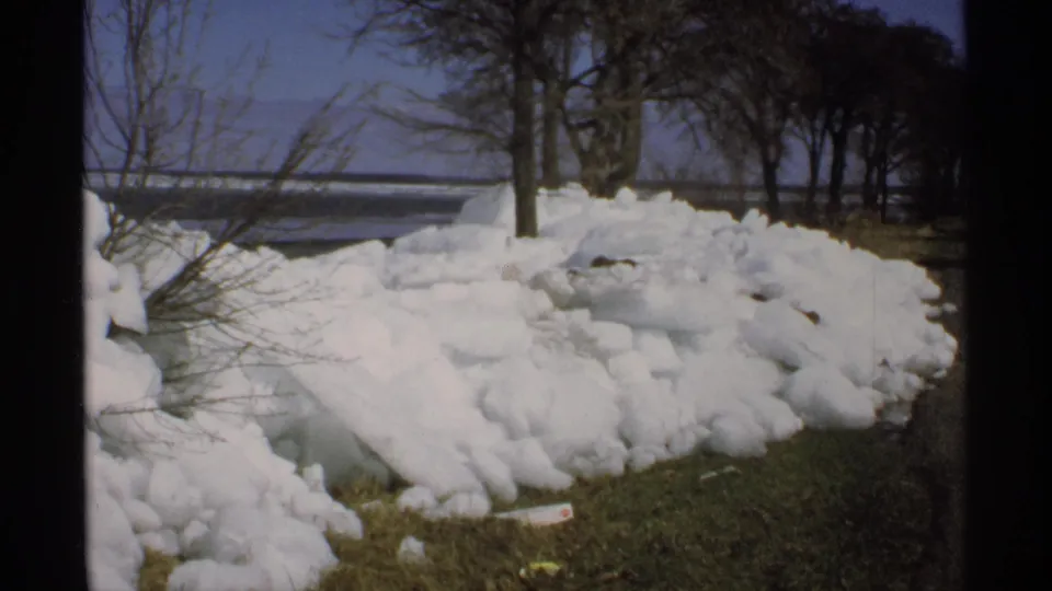 1970 Minnesota 8mm frame showing a large roadside snow pile near grass, bare trees, distant water, and clear sky.
