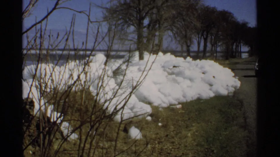 1970 Minnesota home-movie frame with roadside snow, leafless shrubs and trees, a paved road, and water beyond.