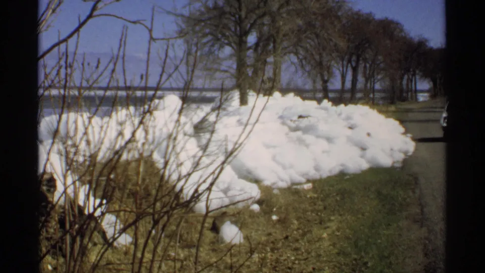 1970 Minnesota 8mm view of a plowed snow mound on grass, with shrubs, roadside pavement, trees, and water in the background.
