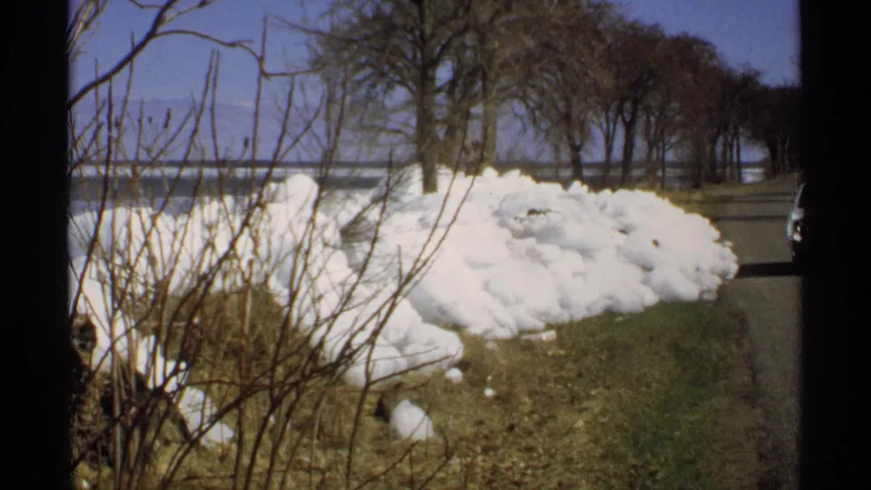 1970 Minnesota frame showing roadside snow, bare trees, distant water, and a partial white vehicle at the right edge.