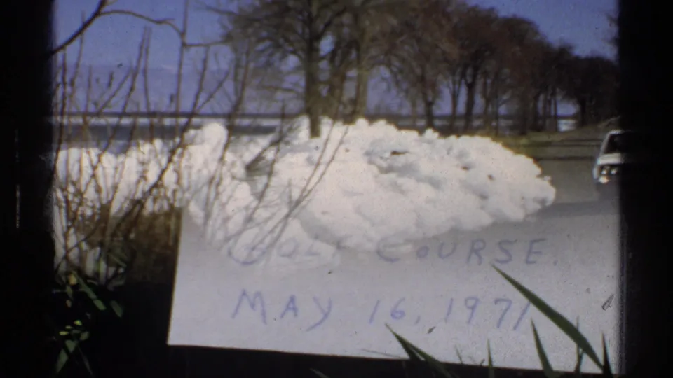 Minnesota 8mm frame showing a snowbank along a roadside, leafless trees, clear sky, and a distant body of water.