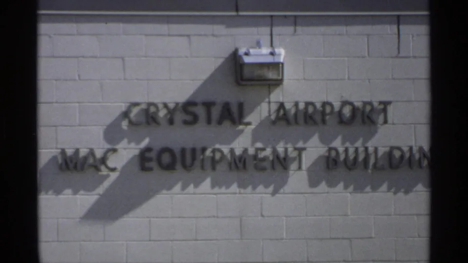Minnesota 8mm frame showing raised lettering for Crystal Airport MAC Equipment Building on a white brick wall with a wall light and shadows.