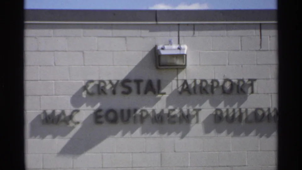 A later 8mm frame of the Crystal Airport MAC Equipment Building sign, with raised letters, a wall light, shadows, and cropped building edge.