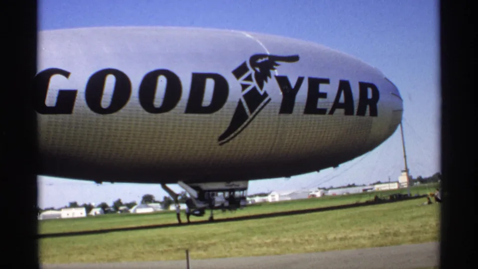 Minnesota 1970 8mm frame of the branded Goodyear blimp envelope, with field, distant structures and airship rigging visible below.