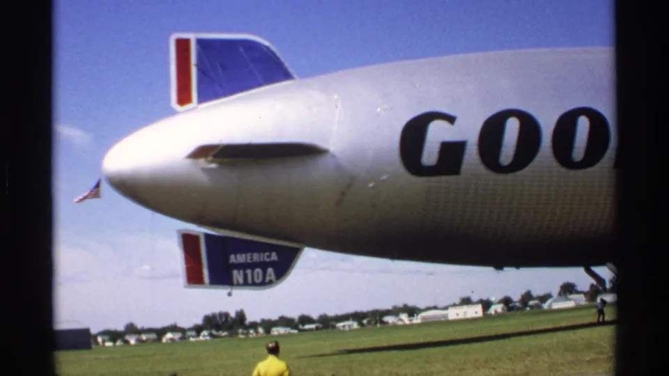Minnesota 1970 8mm frame showing the rear side of the Goodyear blimp, tail fins, partial lettering, a small flag and a person on the field.