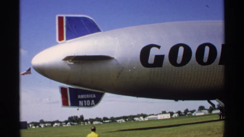 Minnesota 1970 8mm frame showing the Goodyear blimp tail area, partial branded lettering, field, distant buildings and a person at the right edge.