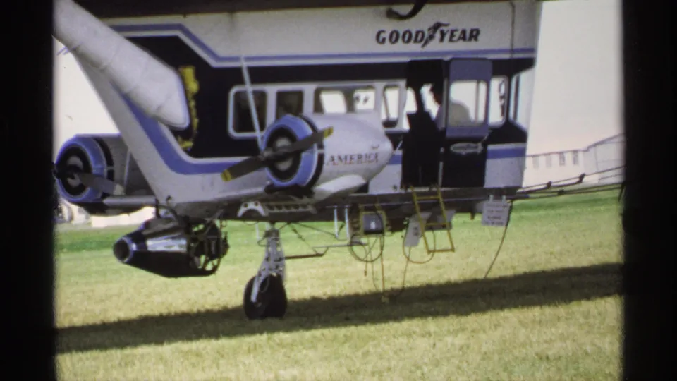1970 Minnesota 8mm frame with a man walking past a Goodyear-branded aircraft section parked on grass.