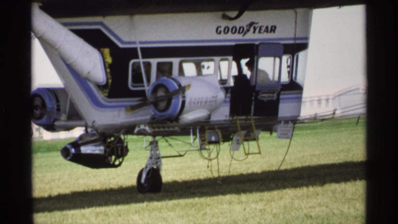 Goodyear blimp parked on grass with propeller engine, ladder, side windows, and open doorway.