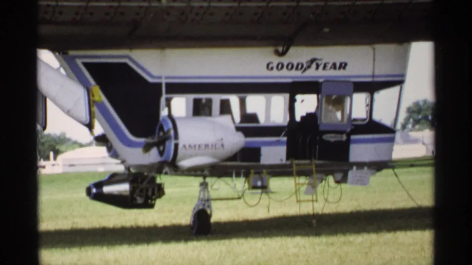 1970 Minnesota frame of the Goodyear blimp on grass, with America lettering, a side ladder, open doorway, and a person visible near the cabin.