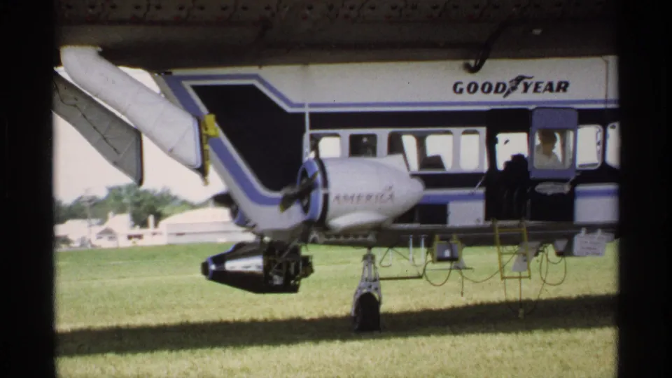 1970 Minnesota 8mm still showing the blimp side, propeller housing, America marking, ladder, open entrance, and grassy field.