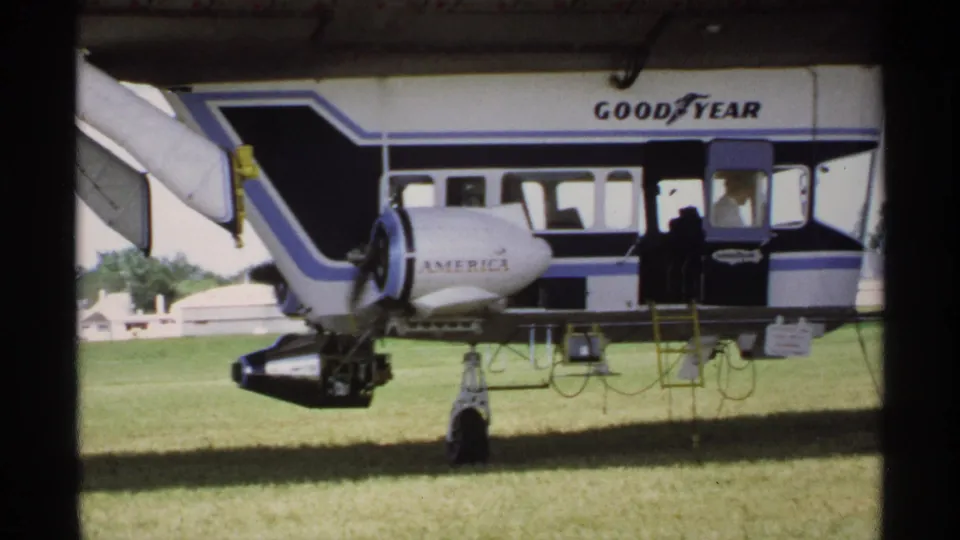 1970 Minnesota frame of a Goodyear blimp parked on a grass field, showing the blue trim, side windows, ladder, engine, and open doorway.