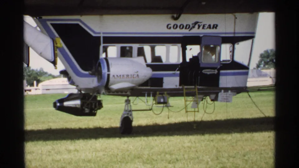 1970 Minnesota 8mm frame of the Goodyear blimp on grass, with the gondola, landing gear, and a person near the doorway.
