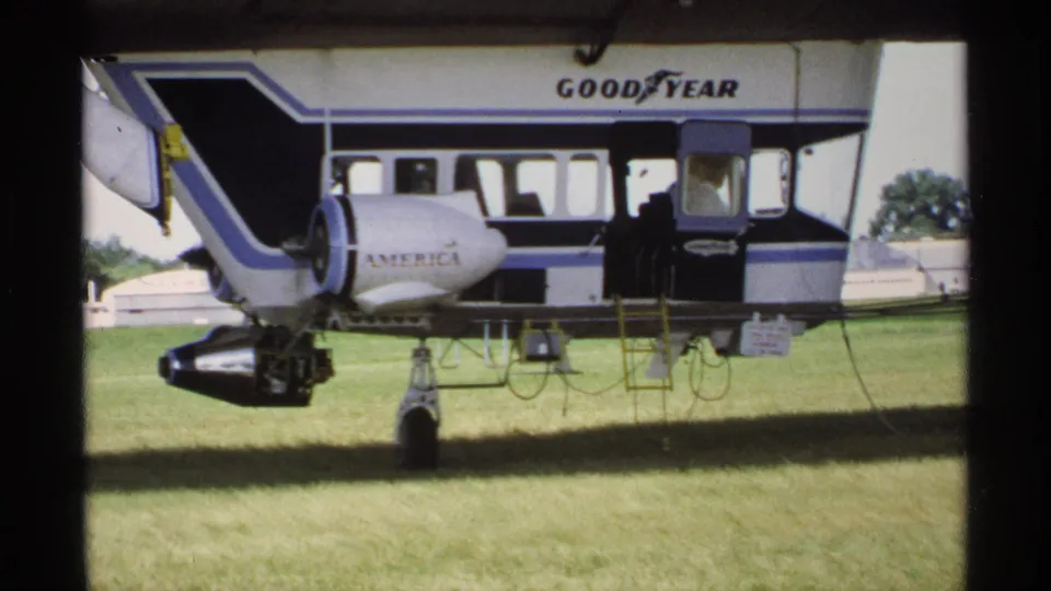1970 Minnesota 8mm frame showing the Goodyear blimp side, AMERICA lettering, gondola area, and landing gear on a sunny grass field.