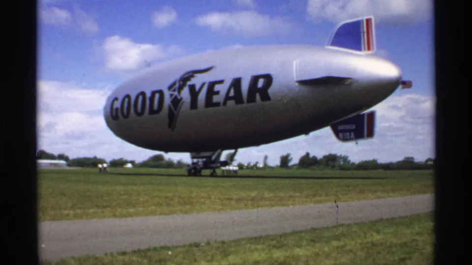 Wider 1970 Minnesota airfield view of the Goodyear blimp America N10A with pavement, grass, and distant figures.