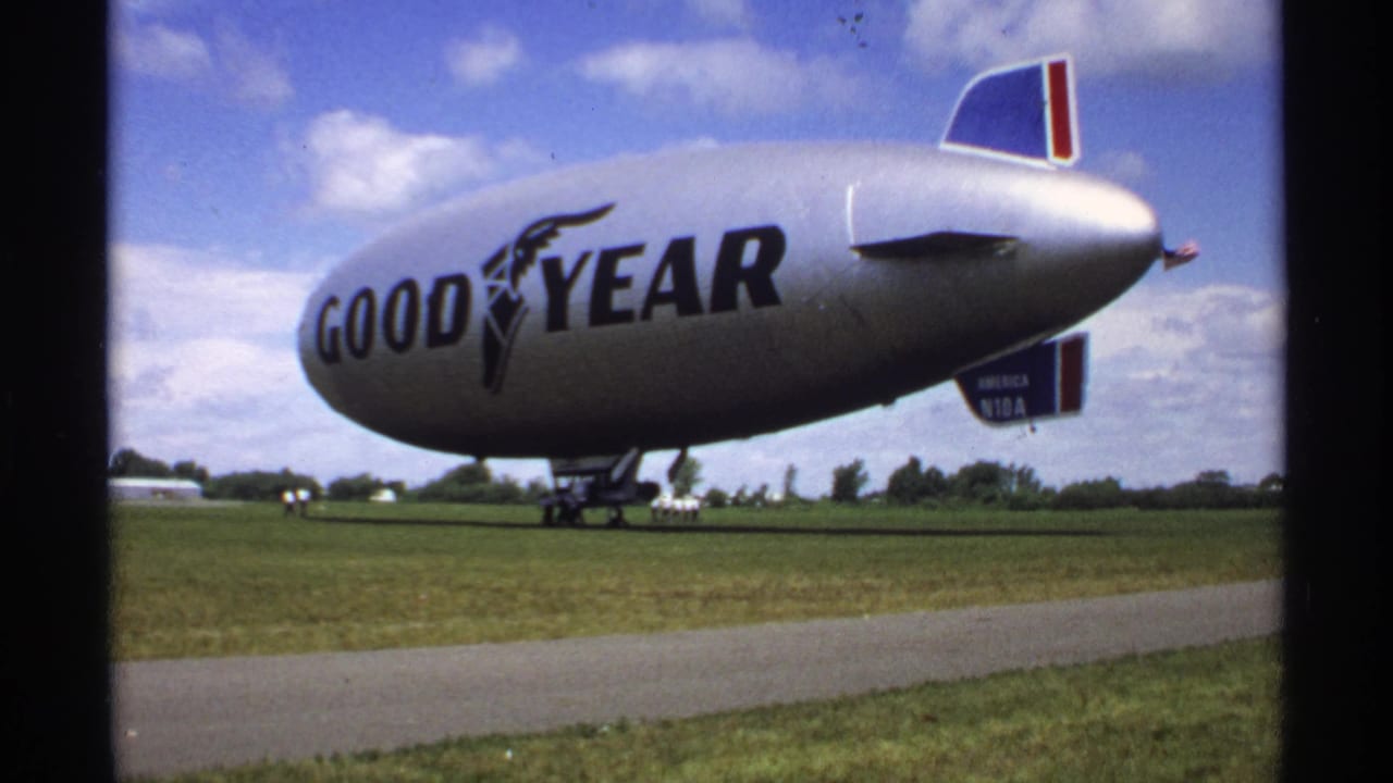 Goodyear blimp resting on grass beneath a blue sky with small figures nearby.