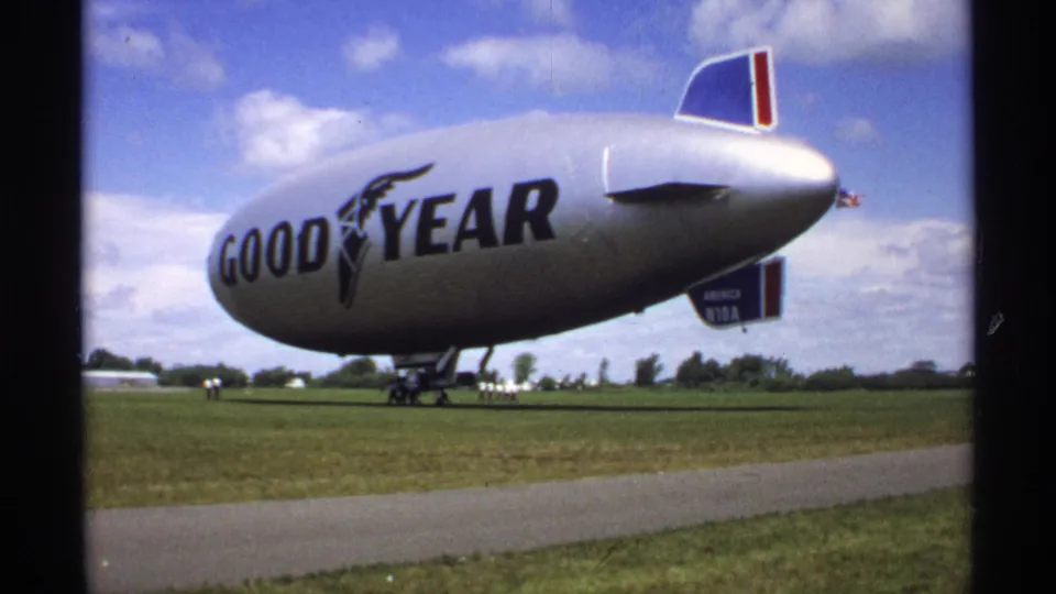 The Goodyear blimp appears grounded on a Minnesota grass field, with landing gear down and small distant people near the airship.