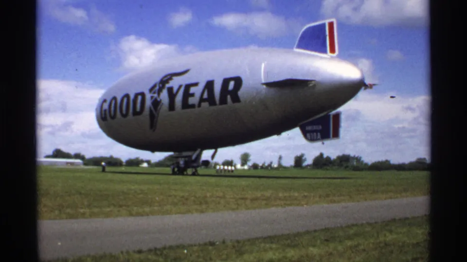 Low-angle 8mm view of the Goodyear blimp on grass, showing the branded envelope, landing gear, tail fins, and open field.