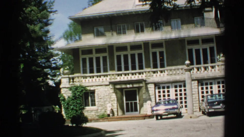 Large Rochester-area building exterior in daylight, with stone lower level, balcony railing, trees, driveway, and two parked cars.