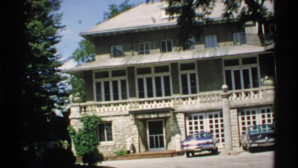 Second exterior view of the large Rochester-area building, showing its balcony, broad facade, shaded trees, driveway, and parked cars.