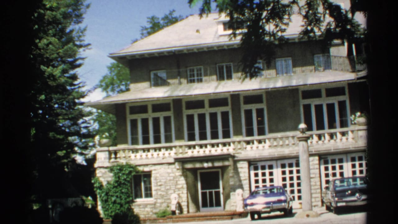 Large residence with stone lower level, balcony, trees, and two cars parked outside.