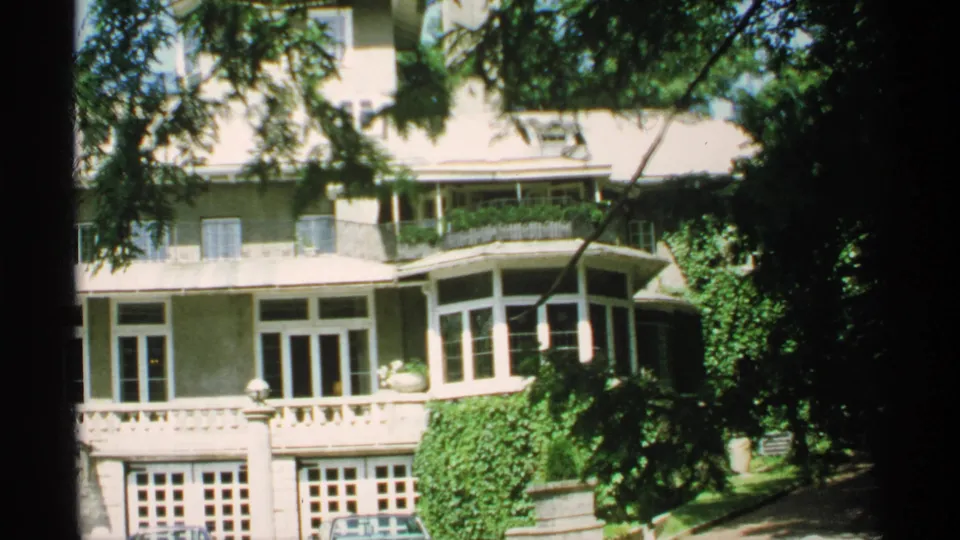 1970 Minnesota home-movie frame with a closer partial view of the residence, balcony doors, ivy growth, shaded trees, and cars below.