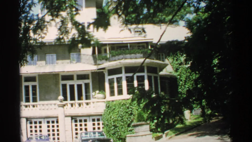 1970 Minnesota 8mm frame showing the residence partly blocked by dark contact-sheet borders, with ivy, balcony railings, trees, and parked cars.