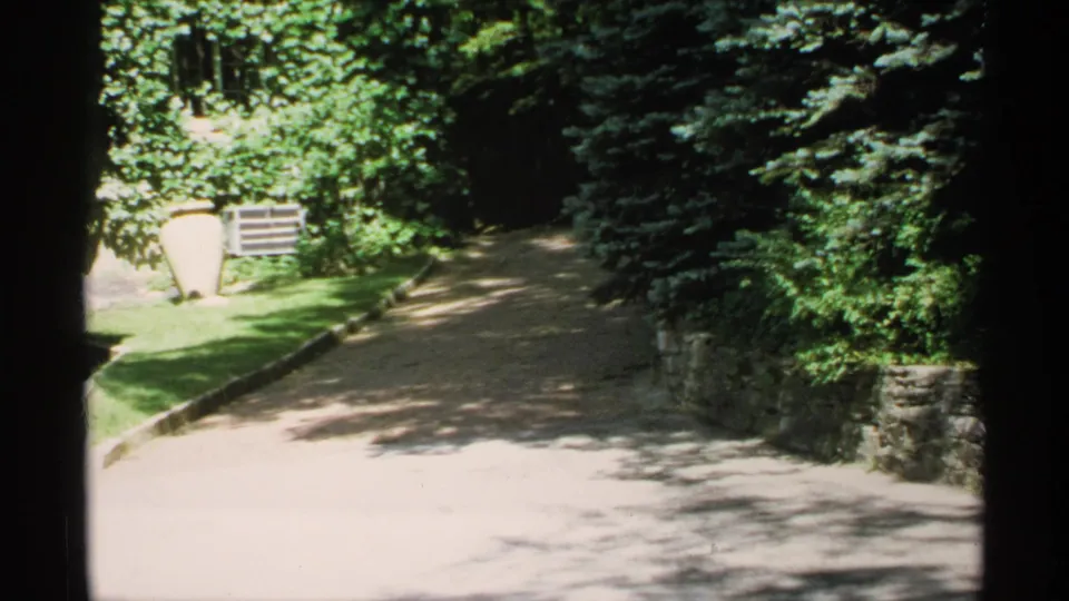 1970 Minnesota 8mm frame of a sun-dappled gravel driveway curving past lawn, dense trees, and a low stone wall.