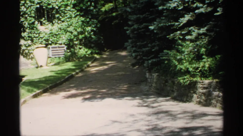 1970 Minnesota home-movie view of a bright gravel drive framed by shrubs, trees, lawn, and stone edging.