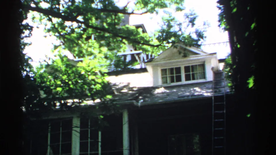 1970 Minnesota 8mm frame of a leafy house exterior with a white-trimmed dormer, shaded porch, and ladder at the side.