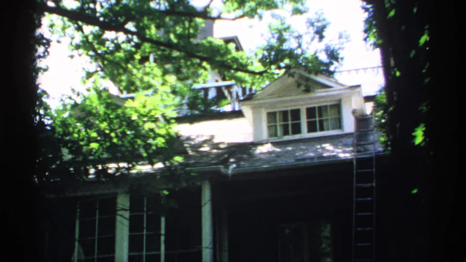 1970 Minnesota 8mm frame showing a shaded residential facade, dormer window, porch columns, tree canopy, and ladder.