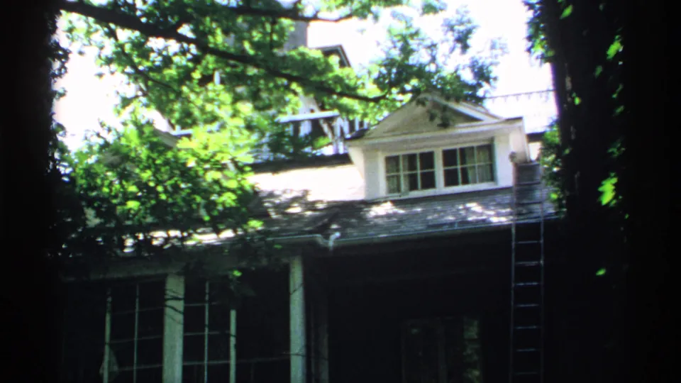 1970 Minnesota 8mm frame of the house exterior from a similar angle, with foliage, white trim, dark porch, and side ladder.