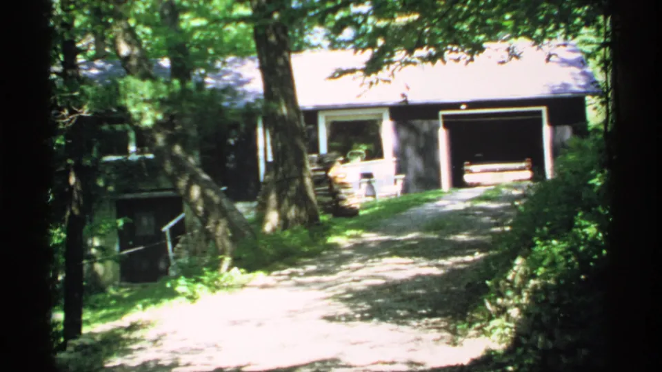 1970 Minnesota 8mm frame of a shaded dirt driveway approaching a modest house with an open garage among trees.