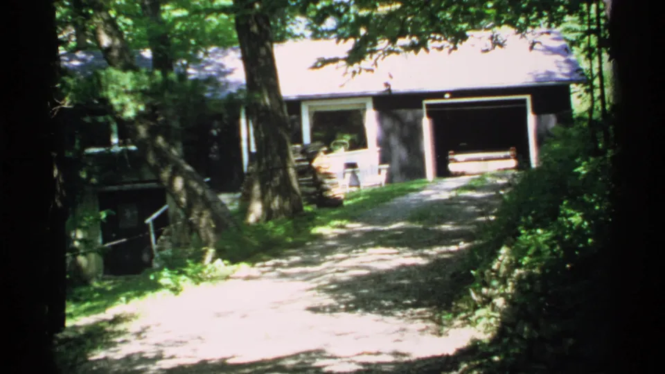 1970 Minnesota home-movie view of a wooded driveway, dark-sided house, and open garage with dappled light.