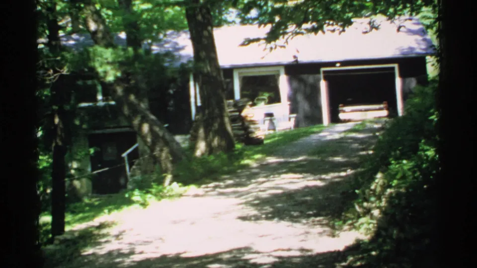 1970 Minnesota 8mm frame showing the same driveway approach to a modest house and open garage under trees.