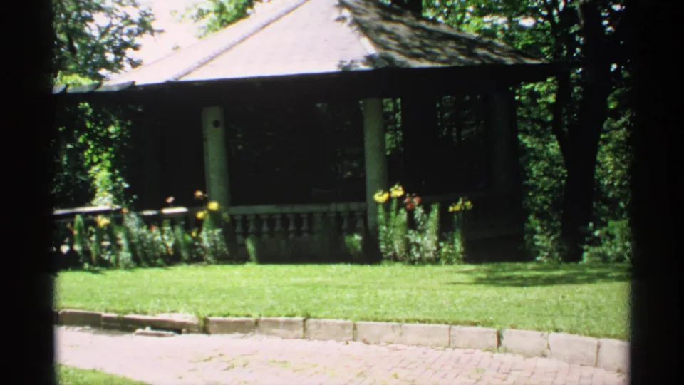 1970 Minnesota home-movie frame of a shaded garden gazebo or porch, manicured lawn, flowers, and low edging.