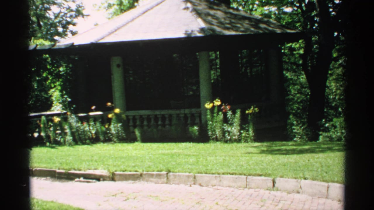 Wooden gazebo behind a green lawn and flower beds in bright garden light.