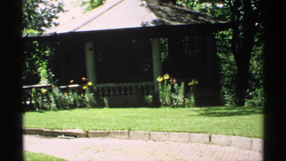 1970 Minnesota garden view with gazebo, shaded trees, flowers, lawn, and bright stone border.