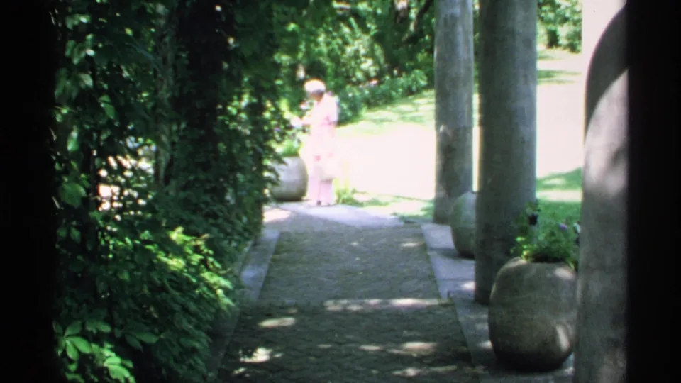 1970 Minnesota 8mm frame of a woman in pink on a sunlit garden path beside columns, planters, and greenery.