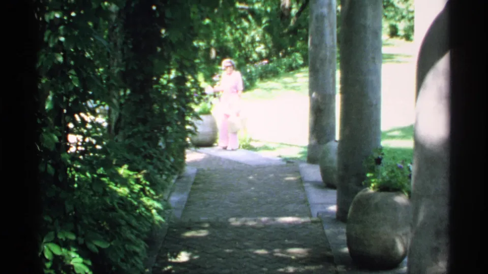 1970 Minnesota garden footage frame with a woman in pink on a bright pathway framed by columns, planters, and foliage.